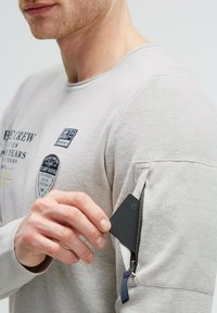 Helles graues Langarmhemd aus strukturiertem Stoff, mit Aufnähern und einer Reißverschlusstasche am Ärmel für sichere Aufbewahrung von Gegenständen.