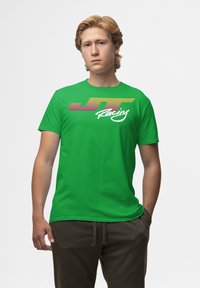 Grünes Baumwoll-T-Shirt mit einem mehrfarbigen "JT Racing"-Grafikdesign in Streifen und in Schreibschrift auf der Vorderseite. Kurze Ärmel und eine lockere Passform.