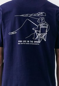 Tricou de culoare albastru marin, cu o ilustratie minimalistă albă care prezintă un urs pescuind dintr-un scaun, cu munți în fundal și text dedesubt.