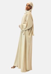 Beige flydende abaya lavet af blødt stof med lange ærmer og en matchende hijab, der har en glat tekstur og et minimalistisk design.