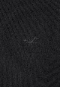 Svart stickad tyg med ribbat mönster. Har en subtil, tonad logotypdetalj som ger en sammanhängande look. Enkelt och minimalistiskt design.