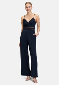 Une combinaison bleu marine avec des bretelles spaghetti fines, un décolleté plongeant, un détail de taille en mesh transparent, des jambes larges et des poches latérales. Des talons hauts argentés.