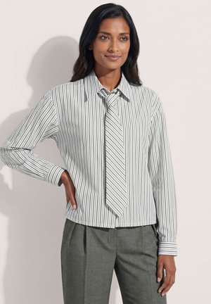Frau trägt ein weiß gestreiftes Langarmshirt mit passender Krawatte und graue Hose, steht mit einer Hand in der Hüfte vor einem einfarbigen Hintergrund.