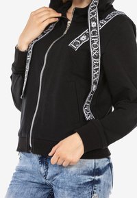 Black zip-up hoodie with patterned white drawstrings, front pockets, and ribbed cuffs. Features a smooth, soft texture.