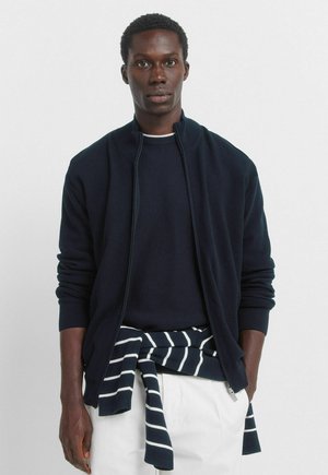 Granatowa kurtka na zamek z prążkowanymi mankietami, noszona na ciemnym swetrze, z pasiastą koszulą zawiązaną w talii. Gładka faktura, lekka tkanina.