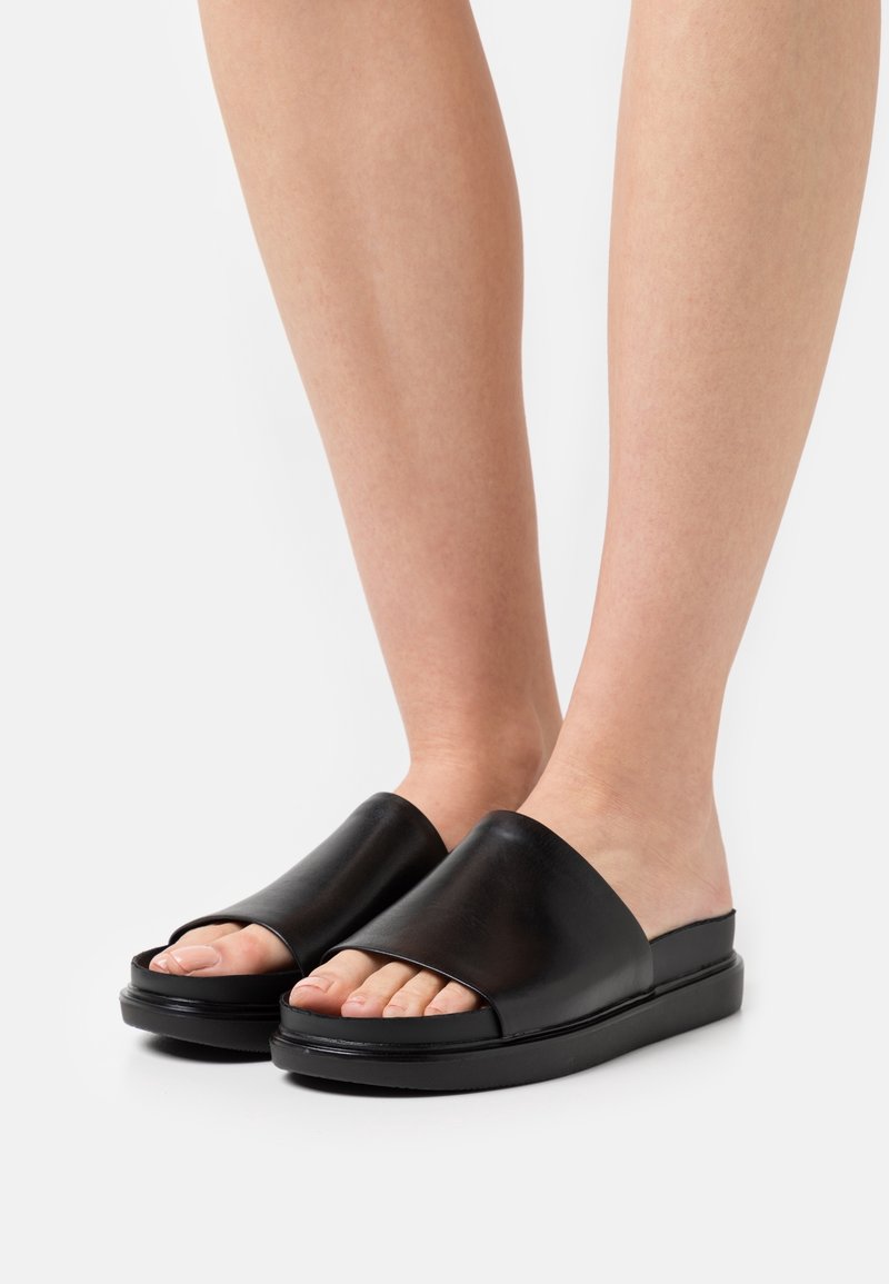 Sandales en cuir noir à glissière avec une large bride supérieure, bout ouvert et semelle plate. Texture lisse avec un matériel minimal et un design épuré.