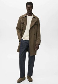 Bruine trenchcoat over een wit overhemd, gecombineerd met donkergrijze broek en bruine schoenen. De jas heeft een dubbelzijdig ontwerp en brede revers.