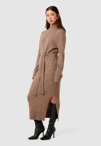 Knielangen, braun gestricktes Kleid mit Gürtel, seitlichen Schlitzen, hohem Kragen und gerippten Bündchen. Kombiniert mit schwarzen High-Heel-Stiefeln.