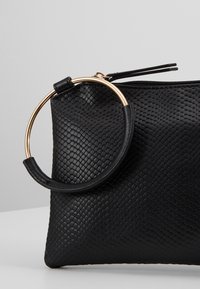 Pochette noire texturée avec une grande lanière de poignet circulaire dorée et noire attachée près de la fermeture éclair.