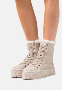 Stivaletti alla caviglia in suede beige con colletto in pelliccia sintetica e suole in gomma spesse e testurizzate. Presentano lacci piatti e occhielli in metallo per la chiusura.