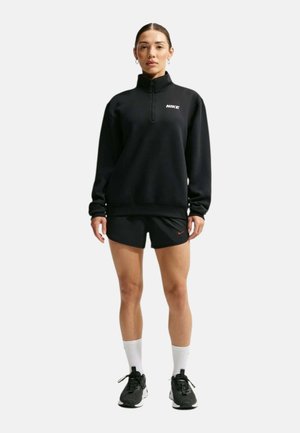 Donna che indossa una felpa Nike nera con zip a quarto di lunghezza, pantaloncini neri, calzini bianchi e scarpe da corsa nere, in piedi su uno sfondo semplice.