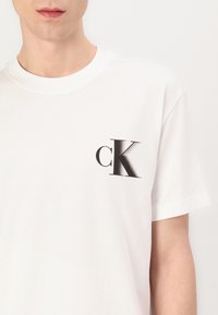White cotton t-shirt featuring a black "CK" logo on the left chest pocket area. Crew neck design with short sleeves and a smooth texture.