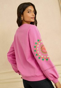 Roze sweatshirt met een ronde hals en pofmouwen. Voorzien van kleurrijke hartvormige borduursels op de linker mouw en een decoratieve zoom.