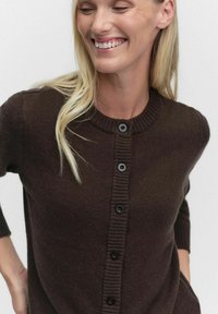 Brun kofta med rund halsringning, ribbade ärmslut och knäppning fram med knappar. Tillverkad av strukturstickad tyg med svarta knappar.