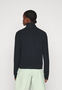 Schwarze Zip-Up-Jacke mit hohem Kragen, langen Ärmeln und flachen Nähten. Das Material wirkt weich und wird mit hellgrünen Shorts kombiniert.