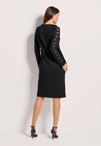 Frau steht mit dem Rücken zur Wand und trägt ein knielanges schwarzes Kleid mit Spitzenärmel-Details und schwarzen High Heels mit Knöchelriemen.