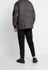 Sort quiltet jakke med høj krave og skrå syninger, parret med sorte bukser og sorte sneakers. Synlig sidelommer.