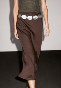 Žena v hnědé saténové midi sukni s širokým kovovým stříbrným diskovým páskem a bezrukávovém hnědém topu s texturou, zobrazena od ramen po lýtka.