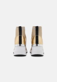 Sneakers alte con tacco metallico dorato, parte superiore in tessuto beige, fascia di accentuazione nera e suola bianca con sottofondo in gomma.