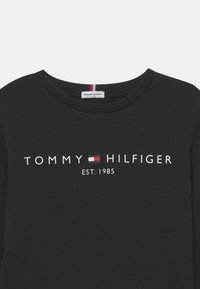 Svart økologisk bomulls T-skjorte med rund hals og hvit "TOMMY HILFIGER EST. 1985" logo, med et lite rødt, hvitt og blått merke på kragen.