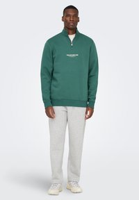 Uomo in piedi che indossa una felpa verde a mezza zip con testo, pantaloni sportivi grigio chiaro e sneaker beige su uno sfondo neutro.