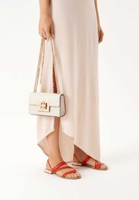 Λευκή τσάντα με χρυσή αγκράφα και αλυσίδα, φορεμένη με μακριά μπεζ φόρεμα. Κόκκινα σανδάλια με λουριά έχουν επίπεδο πάτο και ανοιχτό μπροστινό σχέδιο.