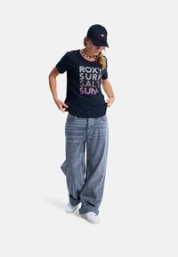 Woman wearing navy cap, shell necklace, navy graphic t-shirt, wide-leg faded jeans, and white sneakers standing with hands adjusting shirt.