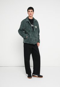 Πράσινο φλις μπουφάν �με υφή, με γιακά και εμπρόσθιες τσέπες, φορεμένο πάνω από μαύρο γραφικό T-shirt και συνδυασμένο με μαύρο παντελόνι και παπούτσια.