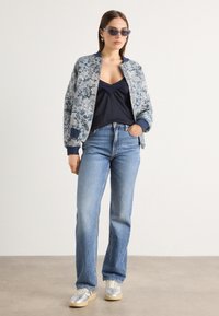 Bomberjack met bloemenpatroon in blauwe tinten, marineblauw shirt, lichtblauwe jeans en zilveren sneakers. Accessoires zijn een zonnebril en oorbellen.
