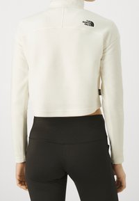 Giacca in pile bianca cropped con colletto alto, caratterizzata da un logo nero sul retro. Abbinata a leggings neri per un look snello.