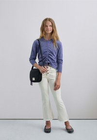 Paarse langlopende top met gathered detail, witte flared jeans, zwarte schoudertas en zwarte flats met witte accenten.