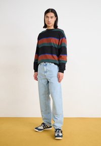 Schwarzer und mehrfarbig gestreifter Sweatshirt; lockere blaue Jeans; dunkle Sneaker mit hellen Akzenten. Das Model steht auf einem gelben Boden vor einem hellen Hintergrund.