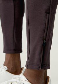 Mörklila träningsbyxor med smal passform, med en dragkedjeficka och mjukt, slätt tygmaterial. Kompletteras av vita sneakers.