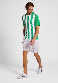 Grön och vit randig idrottströja med korta ärmar, kombinerad med vita shorts. Bärs med vita sportskor med svarta detaljer.