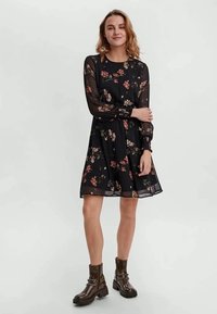 Vestido preto floral com mangas longas transparentes, cintura ajustada e saia em A, apresentando um delicado padrão floral em tons de rosa e verde.