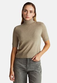 Junge Frau trägt einen beigen kurzärmeligen Rollkragenpullover und graue Jeans und posiert mit einer Hand in der Tasche vor einem weißen Hintergrund.