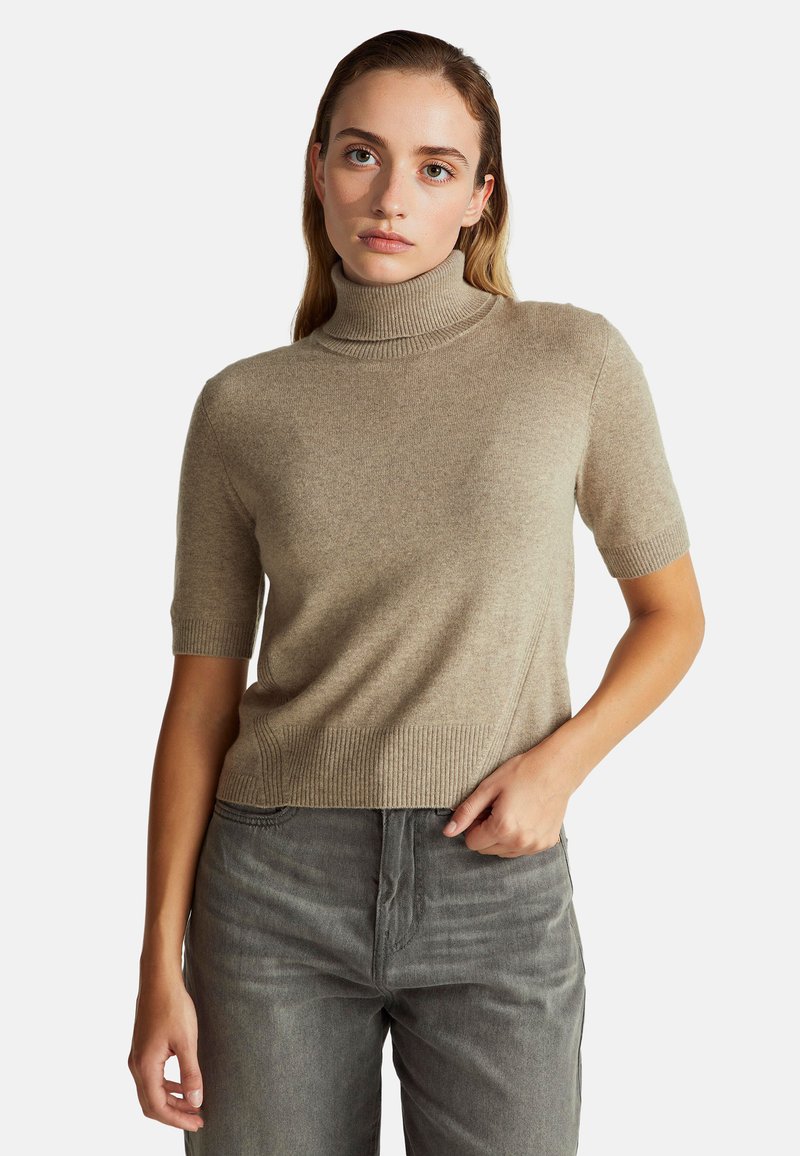 Junge Frau trägt einen beigen kurzärmeligen Rollkragenpullover und graue Jeans und posiert mit einer Hand in der Tasche vor einem weißen Hintergrund.