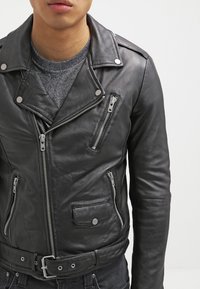 Svart lädermotorcykeljacka med asymmetrisk dragkedja, flera fickor med dragkedja, silverdetaljer och en figursydd midja. Slät yta.