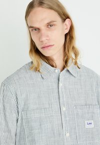 Striped button-up shirt in grey and white cotton with a small pocket and "Lee" label. Features a classic collar and button closure.