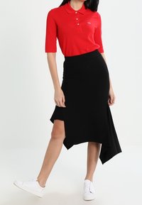 Žena oblečená v červeném polo tričku, černé asymetrické sukni se zářezem a bílých teniskách, stojící na čistém bílém pozadí.