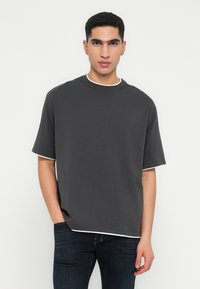 Jonge man met kort krullend haar, gekleed in een oversized donkergrijs T-shirt en donkere spijkerbroeken, staande voor een effen witte achtergrond.
