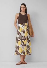 Een bruine tanktop gecombineerd met een witte maxi rok met gele en bruine bladpatronen. Draagt het met tan sandalen en houdt een geweven tas vast.