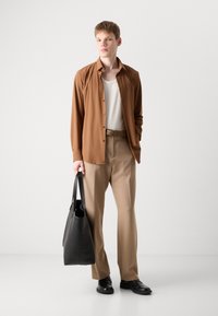Camicia marrone a maniche lunghe sopra una canotta bianca a costine, pantaloni beige, che tiene una borsa a tracolla in pelle nera e indossa scarpe nere.