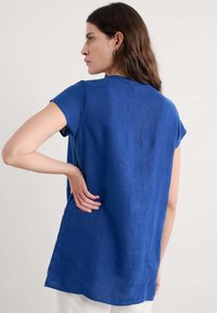 Camisetta blu in lino a maniche corte con vestibilità rilassata, scollatura rotonda e fessure laterali. Il tessuto ha una finitura liscia e leggermente texture.