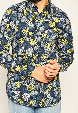 Mand iført en langærmet knap-op-skjorte med et farverigt tropisk bladermønster i blå, gul, hvid og grønne nuancer.