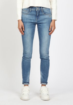 Person, die hellblaue Slim-Fit-Jeans, weiße Turnschuhe und ein weißes Langarmoberteil trägt, vor einem einfarbigen Hintergrund steht.