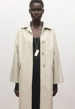 Osoba s tmavou pokožkou nosiaca béžový dlhý kabát cez čierny outfit, doplnená o biely valcovitý prívesok na tenkom dlhom náhrdelníku.