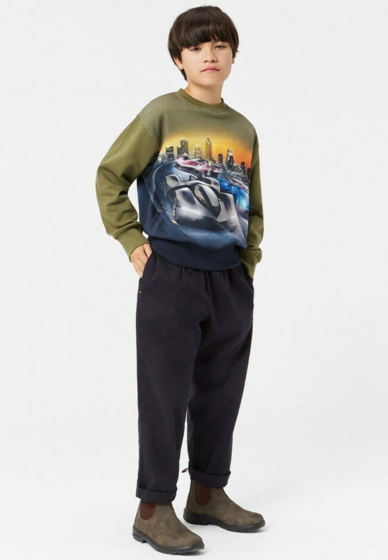 Grafisk sweatshirt med design av en stadssilhuett i gröna och blå nyanser, matchad med mörka, löst sittande byxor och bruna kängor.