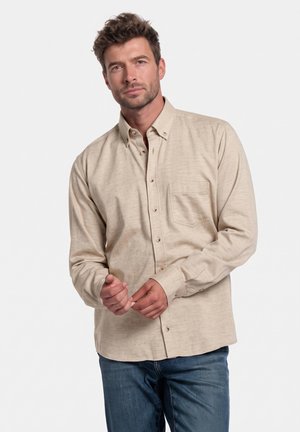 Man met kort krullend bruin haar, gekleed in een beige overhemd met knopen en blauwe spijkerbroek tegen een effen witte achtergrond.