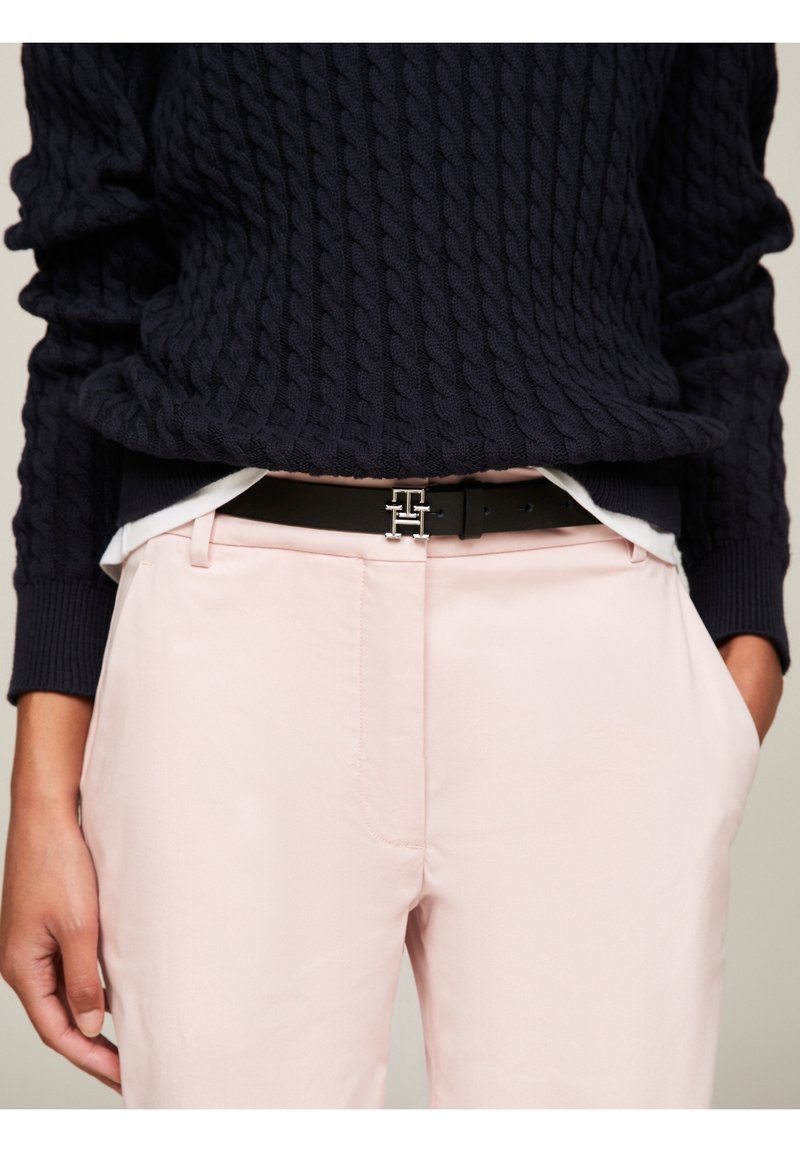 Personne portant un pantalon rose pâle, ceinture noire avec boucle en double T argentée, pull en maille torsadée bleu marine et chemise blanche en dessous.